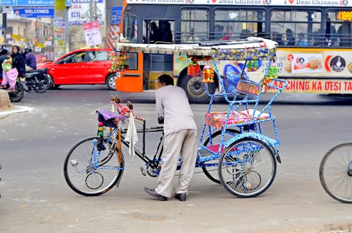 A vibrant rickshaw decorated with colorful ornaments stands on a busy city street. A man is leaning on the rickshaw, which is painted in vivid blues and has intricate patterns. In the background, a crowded bus and a red car can be seen, along with people on motorcycles, indicating a bustling urban environment.