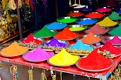 Multiple metal bowls filled with vibrant colored powders are neatly arranged on a table. The colors include red, yellow, green, blue, purple, orange, and pink. Various decorative items are visible in the background, some resembling elephants and traditional Indian ornaments.