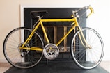 A yellow road bike positioned indoors in front of a black fireplace with a light-colored wall. The bike features a black seat and handlebars, silver rims, and narrow tires. It rests on a dark floor surface.