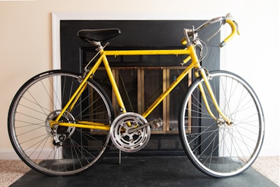 A yellow road bike positioned indoors in front of a black fireplace with a light-colored wall. The bike features a black seat and handlebars, silver rims, and narrow tires. It rests on a dark floor surface.