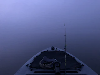 A kayak is positioned in the center with a fishing rod attached, surrounded by a calm and foggy water body. The scene conveys a sense of tranquility and solitude.