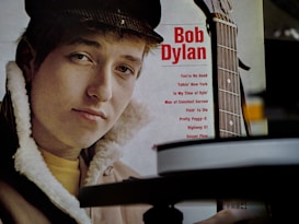 A person is featured prominently, wearing a winter jacket with fur lining and a cap. Next to them, there is a partial view of a guitar neck with text listing songs. The title 'Bob Dylan' is in bold red letters.