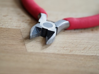 A pair of diagonal cutting pliers with red handles resting on a wooden surface. The metal jaws are slightly open, revealing their sharp edges.
