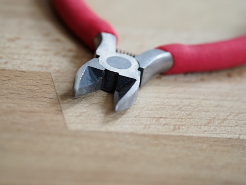 A pair of diagonal cutting pliers with red handles resting on a wooden surface. The metal jaws are slightly open, revealing their sharp edges.