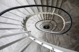 A spiral staircase with a sleek, modern design that spirals downward, creating a mesmerizing optical illusion. The staircase is primarily constructed of gray stone and metal, with a black handrail that contrasts sharply with the lighter steps. The central axis of the spiral reveals a glimpse of greenery at the bottom, adding a touch of life to the architectural scene.