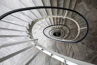 A spiral staircase with a sleek, modern design that spirals downward, creating a mesmerizing optical illusion. The staircase is primarily constructed of gray stone and metal, with a black handrail that contrasts sharply with the lighter steps. The central axis of the spiral reveals a glimpse of greenery at the bottom, adding a touch of life to the architectural scene.