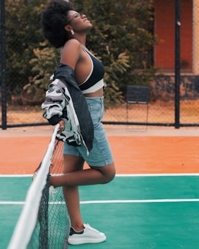 A person stands confidently on a tennis court, leaning on the net with a relaxed posture. They wear a denim skirt, a black and white sports bra, and a jacket draped over the shoulders. White sneakers complement the sporty look. The person gazes upwards with a smile, exuding a carefree and joyful vibe. Lush greenery and a building wall provide a background setting.