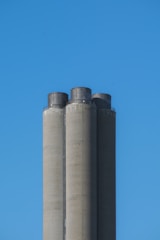 Three tall, cylindrical cement silos stand against a clear blue sky. The structures are slightly weathered, with darker top sections.