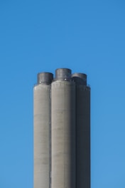 Three tall, cylindrical cement silos stand against a clear blue sky. The structures are slightly weathered, with darker top sections.