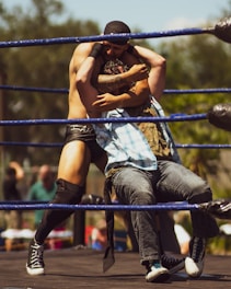 Two individuals are engaged in a wrestling match within a ring surrounded by blue ropes. The person on the left, wearing black gear, has the person on the right in a hold. The latter is dressed in casual attire with a light blue plaid shirt and jeans. The background features blurred trees and a few out-of-focus spectators.