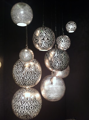 Several intricately designed, spherical pendant lights hang against a dark background. The lights feature detailed patterns with a mix of floral and geometric motifs, casting a warm, diffused glow.