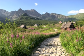 A serene mountainous landscape features traditional wooden huts scattered among lush greenery and vibrant wildflowers. In the foreground, a cobblestone path leads toward the rustic houses, while majestic mountains dominate the background under a clear blue sky.
