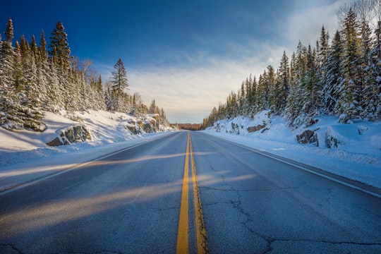 gray asphalt road between snow covered trees under blue sky during daytime in Kirkland Lake Canada