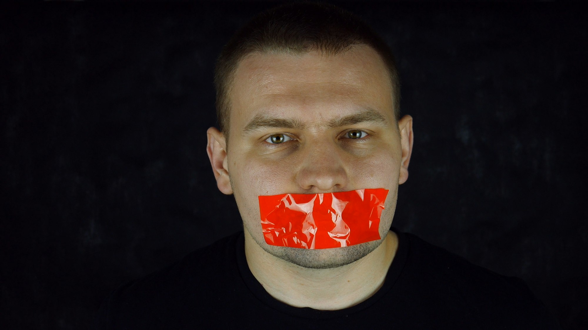 Man with orange scotch-tape on his mouth on black background. Censorship concept