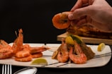 A plate of cooked shrimp is arranged on a white rectangular plate with lemon wedges and herbs. There is a hand holding one of the shrimp. A fork and spoon are visible on the table, and there is another dish in the background.