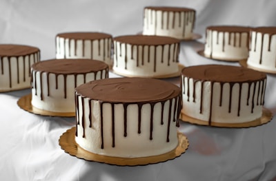 A collection of chocolate-dripped cakes arranged on gold-colored bases. Each cake is decorated with smooth white icing and topped with a glossy chocolate ganache that drips down the sides.