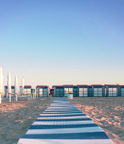 A sandy beach with a patterned walkway leading to a row of beach cabins. Each cabin has a blue and white exterior with red roofs. On the left, closed outdoor umbrellas stand on a deck area adjacent to the cabins. The sky is clear and blue, suggesting a calm day at the beach.