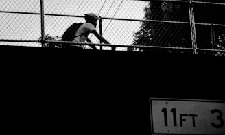 A person wearing a backpack and helmet rides a bicycle behind a chain-link fence, with a road sign showing a clearance height of 11 feet 3 inches visible in the foreground. The image is presented in high contrast black and white, creating a strong silhouette effect.