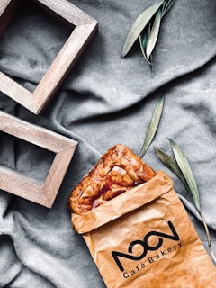 A brown paper bag from a cafe bakery contains a golden-brown pastry. The scene is set on a textured gray fabric, with green leaves and wooden rectangular frames adding a stylish touch.