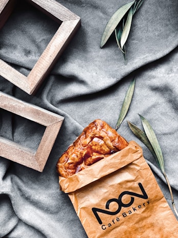 A brown paper bag from a cafe bakery contains a golden-brown pastry. The scene is set on a textured gray fabric, with green leaves and wooden rectangular frames adding a stylish touch.