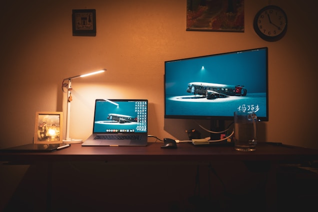 A dimly lit workspace featuring a laptop and a larger monitor displaying the same underwater wreckage image. To the left, a desk lamp casts a warm glow over a framed picture, and there's a clock on the wall. Various objects like a mug, a stylus pen, and cables are scattered on the desk, creating a homely and functional atmosphere.
