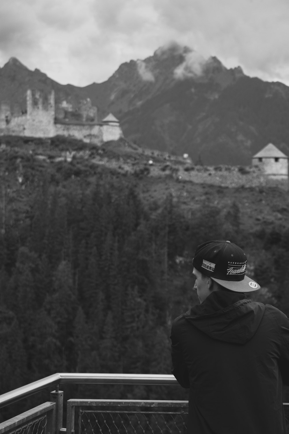 grayscale photo of man in jacket and cap looking at the mountains
