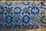 The image consists of a wall covered with detailed blue and white ceramic tiles. The tiles feature intricate floral and geometric patterns, with some visible signs of wear and chipping, revealing a rustic texture. Shadows cast across the tiles suggest natural light illuminating the surface, highlighting the vibrant blue hues.