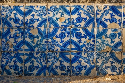 The image consists of a wall covered with detailed blue and white ceramic tiles. The tiles feature intricate floral and geometric patterns, with some visible signs of wear and chipping, revealing a rustic texture. Shadows cast across the tiles suggest natural light illuminating the surface, highlighting the vibrant blue hues.