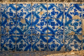 The image consists of a wall covered with detailed blue and white ceramic tiles. The tiles feature intricate floral and geometric patterns, with some visible signs of wear and chipping, revealing a rustic texture. Shadows cast across the tiles suggest natural light illuminating the surface, highlighting the vibrant blue hues.