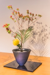 A minimalist floral arrangement is presented in a sleek, dark vase set on a reflective surface. The arrangement includes delicate branches with small red and yellow flowers, complemented by a bright yellow spherical bloom and green leaves. The background is softly lit, casting gentle shadows that enhance the overall composition.