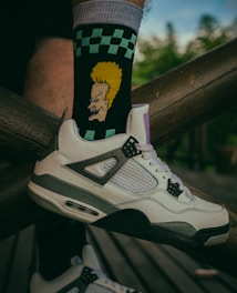 A close-up of a person's foot wearing a white and gray sneaker, paired with a black sock featuring a cartoon character with yellow hair. The sock also has a green and black checkerboard pattern. The foot is resting on a wooden structure with a blurred green background.