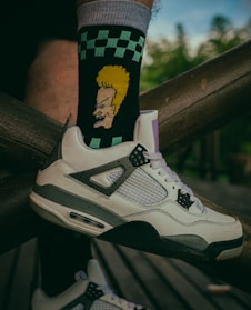 A close-up of a person's foot wearing a white and gray sneaker, paired with a black sock featuring a cartoon character with yellow hair. The sock also has a green and black checkerboard pattern. The foot is resting on a wooden structure with a blurred green background.