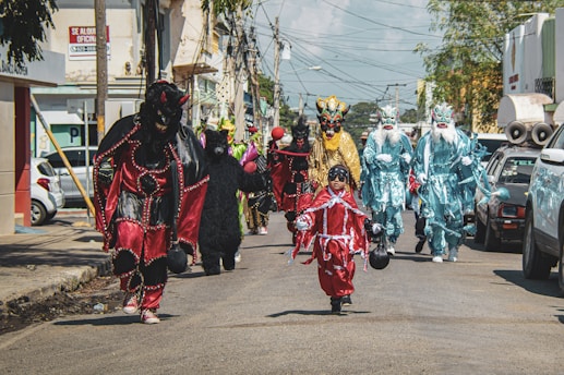 A group of people dressed in elaborate and colorful costumes, including masks and robes, are walking down a street. The costumes resemble fantastic creatures or mythical beings, with vibrant reds, blues, and yellows. The scene captures a festive or cultural event, likely a parade or celebration.