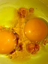 Two raw egg yolks surrounded by egg whites with many air bubbles visible, set against a yellow background. The yolks are vibrant and glossy, with the bubbles creating an interesting texture.