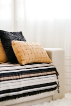 A cozy couch with a striped blanket in shades of black, brown, and cream is adorned with two textured pillows, one in a woven tan fabric and the other in a soft black material. Sunlight filters softly through sheer curtains, casting a warm and inviting atmosphere.