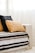 A cozy couch with a striped blanket in shades of black, brown, and cream is adorned with two textured pillows, one in a woven tan fabric and the other in a soft black material. Sunlight filters softly through sheer curtains, casting a warm and inviting atmosphere.