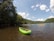 A bright green kayak rests on the shallow waters of a river, surrounded by lush green forest. The sky is partly cloudy with patches of blue visible. Several kayakers are seen paddling in the river, using colorful kayaks in shades of yellow, orange, and blue.