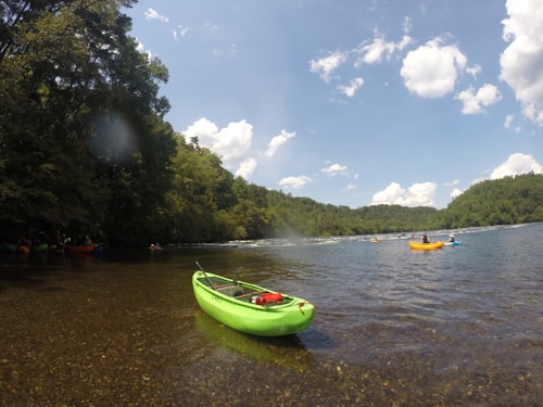 A bright green kayak rests on the shallow waters of a river, surrounded by lush green forest. The sky is partly cloudy with patches of blue visible. Several kayakers are seen paddling in the river, using colorful kayaks in shades of yellow, orange, and blue.