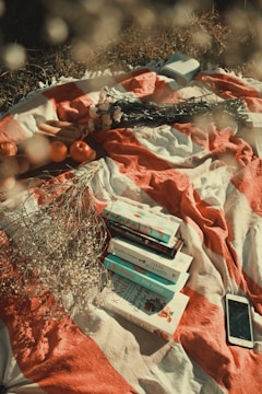 A cozy picnic setup featuring a striped blanket with several books neatly stacked. Scattered around are delicate white flowers and two oranges. A woven straw hat and a smartphone are also visible, along with a small portable speaker.