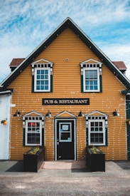 A charming yellow building with white trim labeled 'Pub & Restaurant' has two front windows with geometric window panels. An 'Open' neon sign is displayed in one of the windows. Black planters with small plants are positioned on either side of the entrance, and three vintage-style black lamps are mounted above the windows and door. The building sits against a backdrop of a partly cloudy sky.