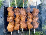 Several skewers are lined up side by side, each loaded with chunks of marinated meat being grilled over an open flame. The meat appears juicy and well-seasoned, with a charred appearance on the edges, indicating it is cooking over a high heat. Smoke wafts up from the grill, suggesting the barbecue is in full swing, while greenery and grass are visible in the background.