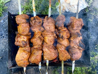 Several skewers are lined up side by side, each loaded with chunks of marinated meat being grilled over an open flame. The meat appears juicy and well-seasoned, with a charred appearance on the edges, indicating it is cooking over a high heat. Smoke wafts up from the grill, suggesting the barbecue is in full swing, while greenery and grass are visible in the background.