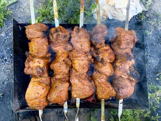 Several skewers are lined up side by side, each loaded with chunks of marinated meat being grilled over an open flame. The meat appears juicy and well-seasoned, with a charred appearance on the edges, indicating it is cooking over a high heat. Smoke wafts up from the grill, suggesting the barbecue is in full swing, while greenery and grass are visible in the background.
