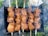 Several skewers are lined up side by side, each loaded with chunks of marinated meat being grilled over an open flame. The meat appears juicy and well-seasoned, with a charred appearance on the edges, indicating it is cooking over a high heat. Smoke wafts up from the grill, suggesting the barbecue is in full swing, while greenery and grass are visible in the background.