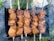 Several skewers are lined up side by side, each loaded with chunks of marinated meat being grilled over an open flame. The meat appears juicy and well-seasoned, with a charred appearance on the edges, indicating it is cooking over a high heat. Smoke wafts up from the grill, suggesting the barbecue is in full swing, while greenery and grass are visible in the background.