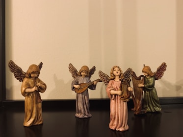 Four small angel figurines are arranged in a line, each holding a different musical instrument: a flute, a lute, a harp, and possibly a violin. They have detailed wings and are wearing robes of various colors including brown, purple, pink, and green. The base they are standing on and the background is plain.