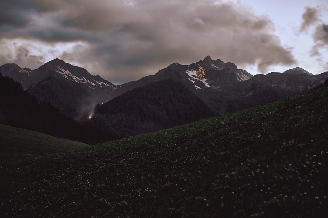 green grass field near snow covered mountain under cloudy sky during daytime