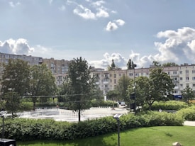 A cityscape featuring a cluster of mid-rise apartment buildings with a foreground of lush greenery, trees, and a small fountain. The sky is partly cloudy with patches of blue showing through.