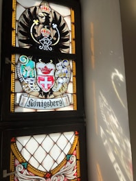 A stained glass window featuring a coat of arms with an eagle and a crown motif. Below it, there's ornate heraldry with shields and crests, including symbols such as a cross and another crown. The word 'Königsberg' is inscribed at the bottom, and light reflects off the window onto the adjacent wall.