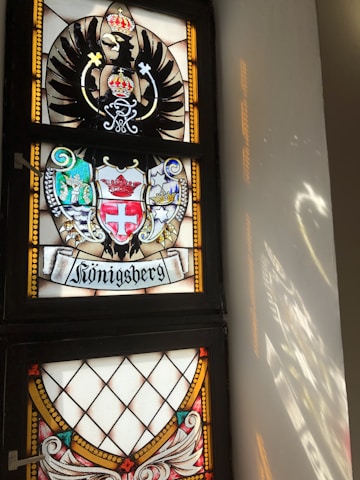 A stained glass window featuring a coat of arms with an eagle and a crown motif. Below it, there's ornate heraldry with shields and crests, including symbols such as a cross and another crown. The word 'Königsberg' is inscribed at the bottom, and light reflects off the window onto the adjacent wall.